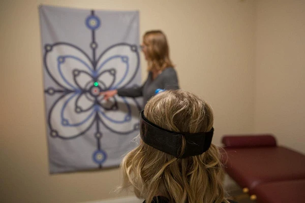 A patient wearing a headband device participates in a visual or neurological assessment, focusing on a patterned wall chart with a laser pointer.